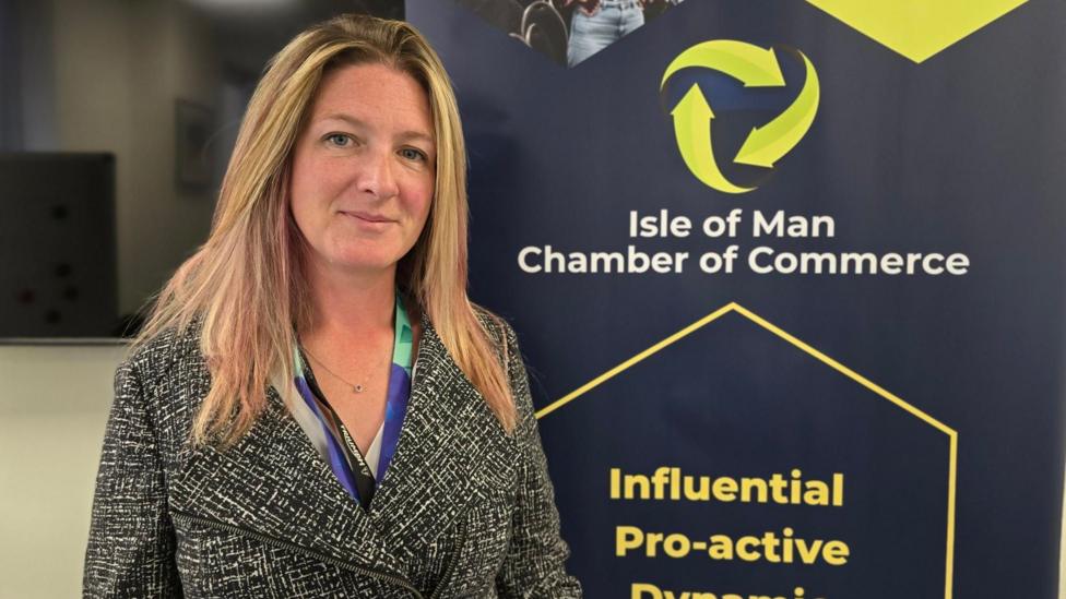 Claire has long straight blonde hair with light pink highlights coming through. She is wearing a black and white patterned suit jacket and standing next to a large navy Isle of Man Chamber of Commerce banner.