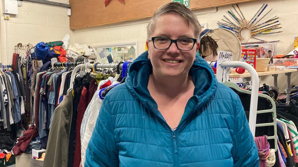 Katie Wilson wearing glasses and a blue coat with a blue hood smiling at the camera. Behind here is racks full of different clothes.