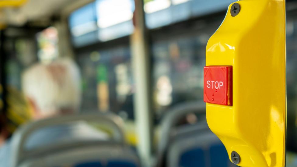 A close up of a red "stop" button which passengers press on the bus to alert the driver they want to get off at the next stop. The front of the bus is out of focus but we can make out the head and shoulders of an older white-haired man.