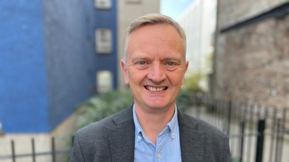 Paul White, a middle-aged man with greying hair, smiles against the backdrop of a blue painted building. He wears a pale blue shirt and a dark grey suit jacket.