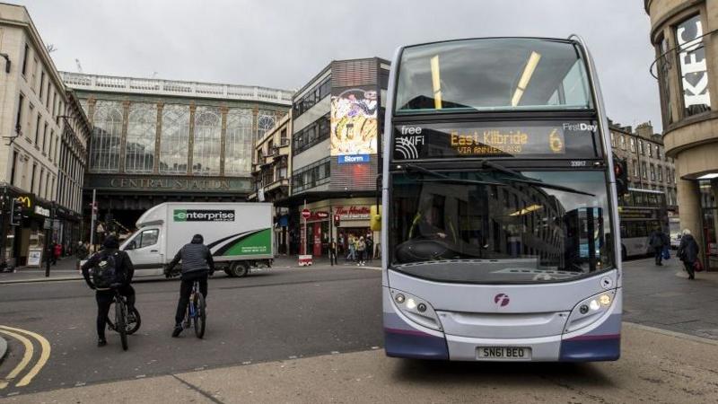 At the junction of four roads in Glasgow, a white double decker bus swings around the corner into shot, giving the impression it is coming right at us. Its sign reads "East Kilbride, via Anniesland and the number 6. Two cyclists wait to go the opposite way and people cross the road to the other side of the bus. Behind we see a lit up advertising screen and a McDonalds in the distance.