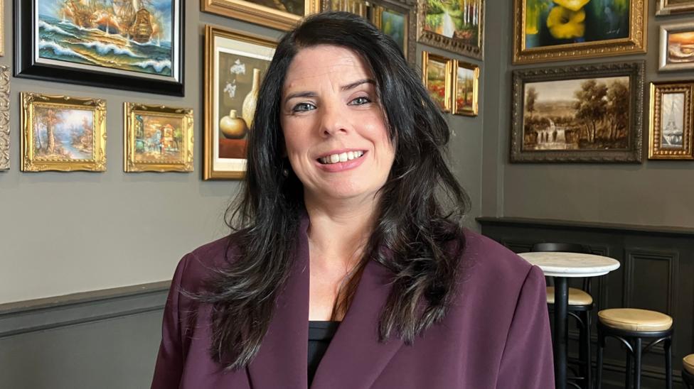 A smiling Anne Marie Gallagher is wearing a crimson jacket. She has long dark hair and is standing in a room in which the walls are covered in paintings. A table and chairs are in the background