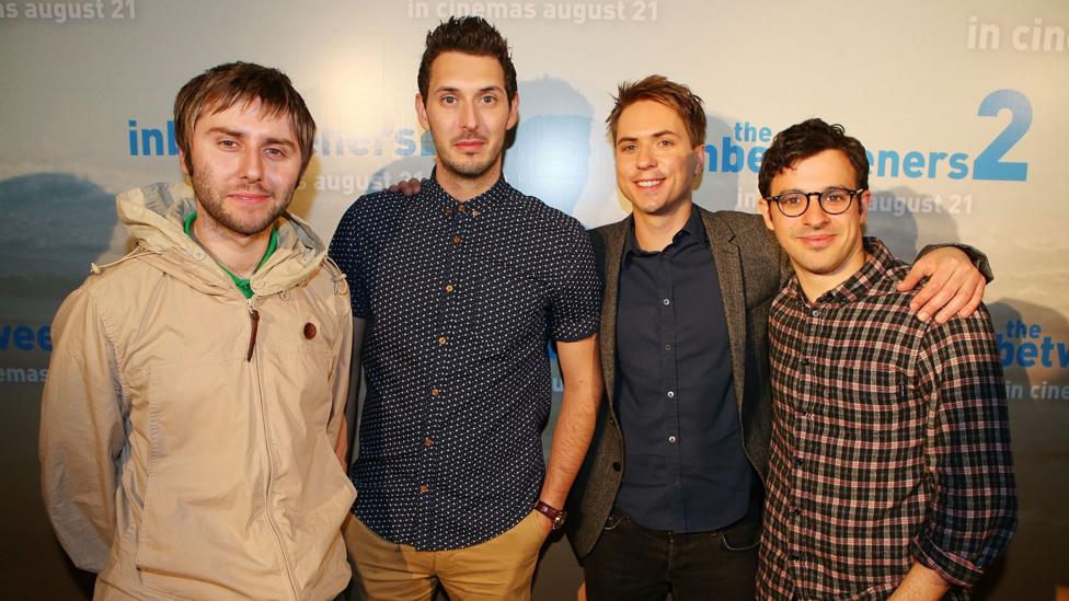 James Buckley, Blake Harrison, Joe Thomas and Simon Bird pose at a premier of The Inbetweeners 2 in front of a billboard of the film. James wears a grey waterproof, Blake blue and white patterned top, Joe wears a grey blazer over a navy shirt and Simon wears checked patterned shirt.