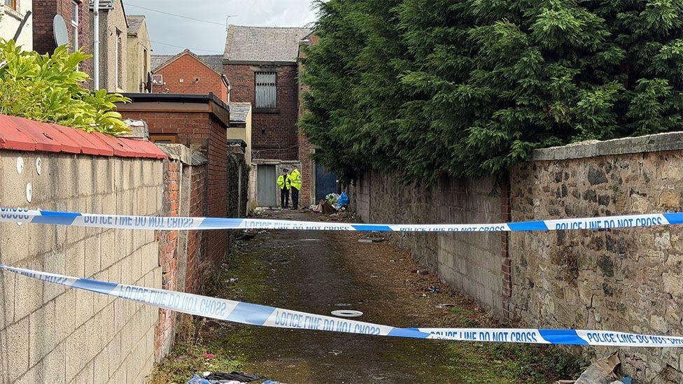 Police cordon tape blocks access to a narrow alleyway between brick and stone walls, with two high-visibility jacketed officers visible in the distance.