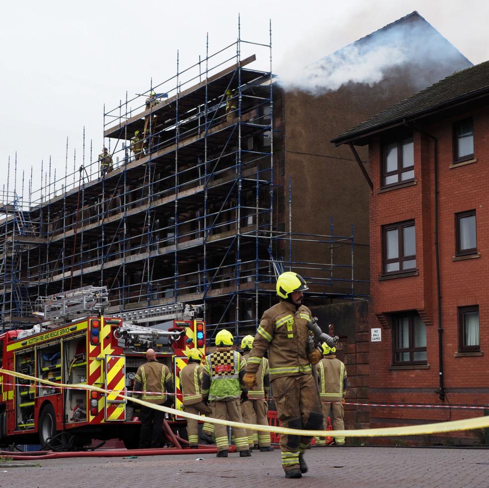 Fie engine and a number of firefighters in a cordoned-off area of a road. There is snoke coming from the roof of the building which is covered by scaffolding.