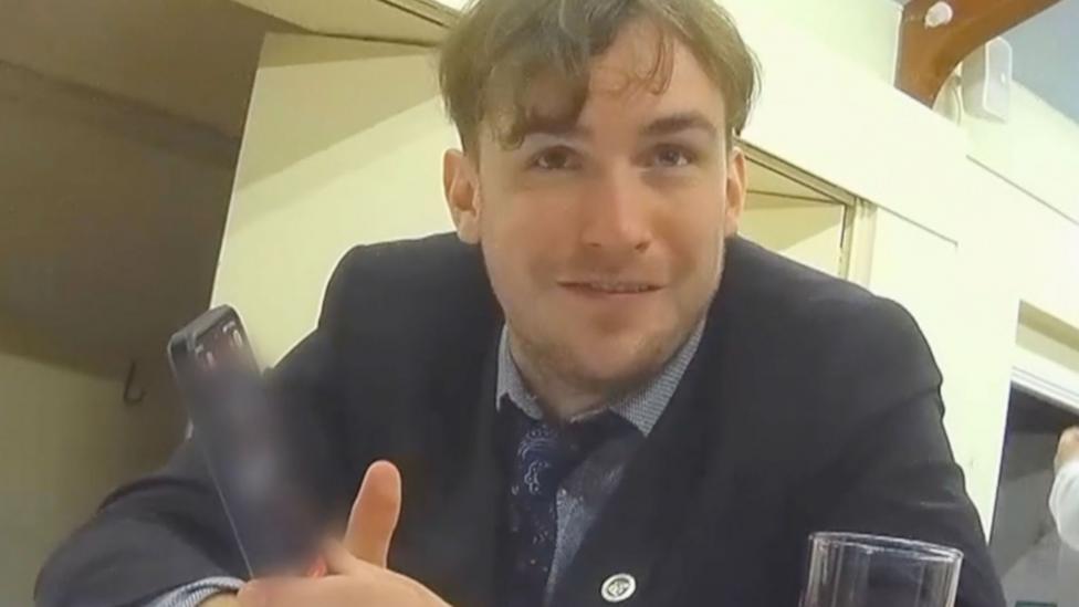 Webster, who is wearing a suit, holding his phone; the screen has been blurred