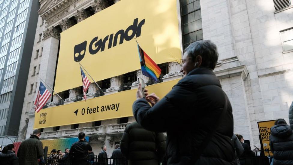 A woman talks to someone on a video call on her smartphone in front of the building housing the New York Stock Exchange on Wall Street, which is covered in a huge Grindr poster to mark the company going public in November 2022. A LGBTQ+ flag is flying beneath it, alongside the US flag, with people milling around below.