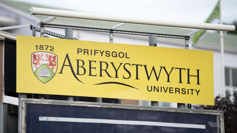 A sign for Aberystwyth University. It is a metal sign with a yellow background, with the text in black. There is also a logo for the university on the sign, which is a shield featuring two dragons in the top half, and an open book in the bottom half.