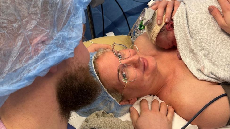 A photo of woman holding her new born baby after giving birth, with the back of her partners head in scrubs.