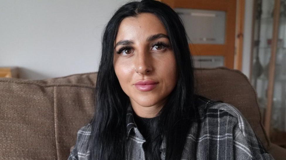 A woman with long black hair wearing a tartan shirt, sitting on the sofa.