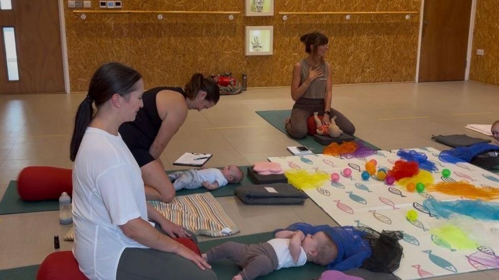 Mother and baby yoga with an instructor sitting in front of the class and two mothers and their babies following instructions.