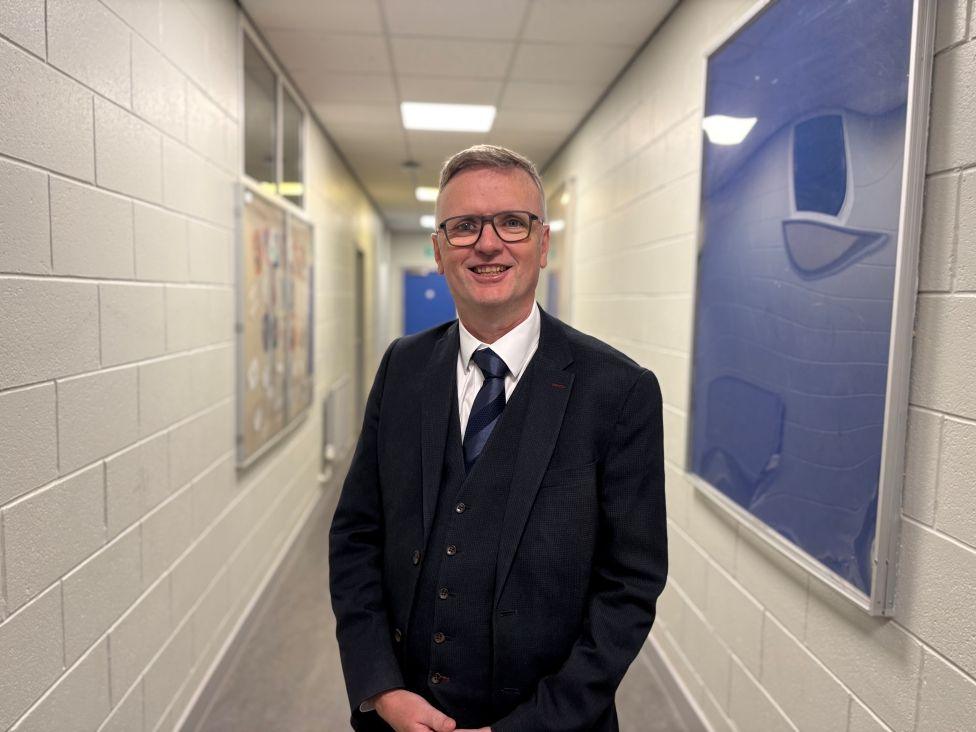 A man with glasses in a dark suit stands in a bright school corridor with white walls