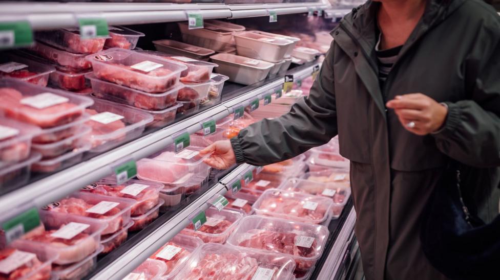 A woman wearing a long khaki coat reaches into a supermarket fridge filled with packaged meat.
