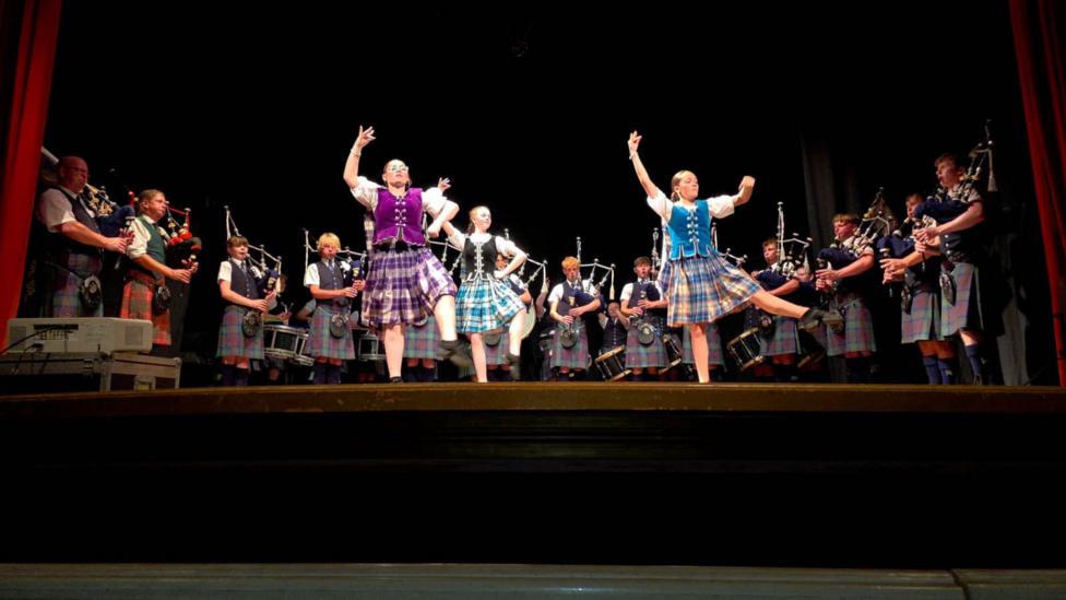 Traditional Highland dancers on stage with a pipe band behind them
