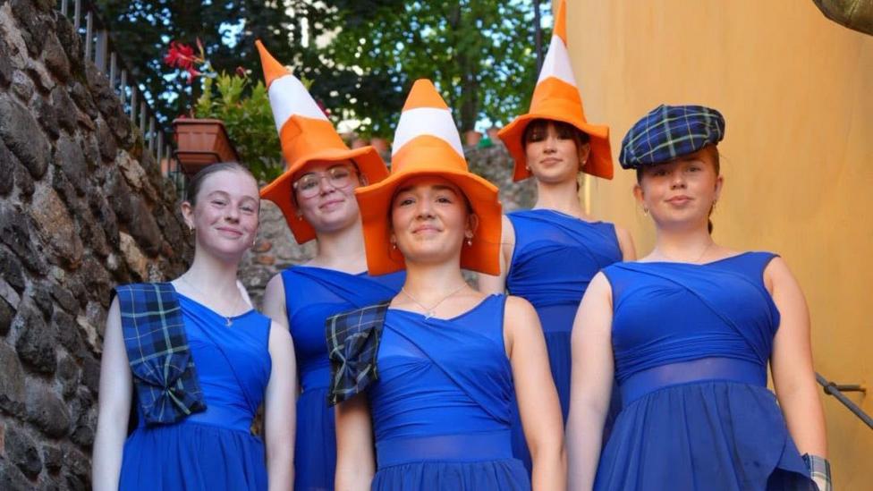 A group of young female dancers, three of them with traffic cone hats on their heads and another with a tartan bunnet