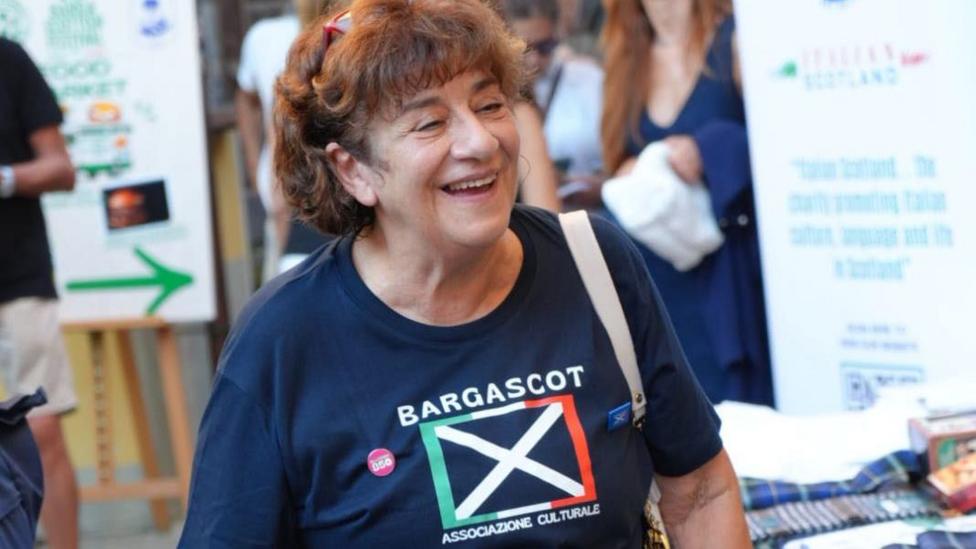 A woman in a BargaScot T-shirt smiles. She has curly brown hair and her glasses up on her head.