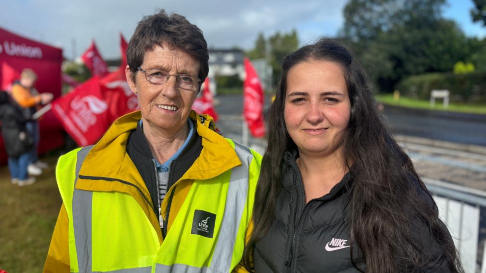 Two women stand side by side along a main road. The woman on the left is older, with short hair and is wearing a high visibility jacket. Beside her is a woman in a black coat, with long dark hair. Both are smiling at the camera. Behind them is a gathering of workers on strike. they are brandishing flags with their unions name and waving at passing cars.