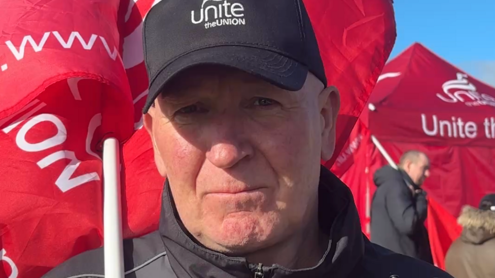 A man is looking at the camera with a serious expression. He's wearing a black baseball cap that has cast a shadow over his face. He has a red flag on a flagpole hanging over his shoulder. and behind him billow more of the bright red unite flags.