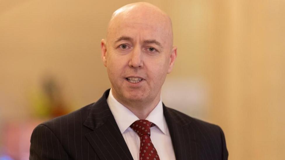A bald man looks into the camera. He is wearing a black suit jacket, white shirt and red tie.