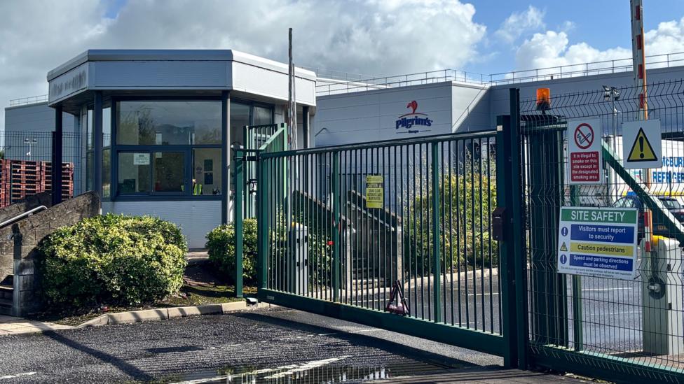A building can be seen behind a closed gate. The logo Pilgrim's can be seen on the building. beside the high, green, metal gate is a security hut. It's painted grey and has windows.