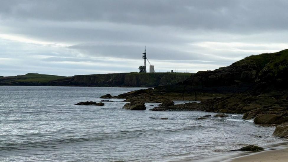 A different angle of the spaceport, seen this time from the beach. In the distance, a tall launch pad is on a long flat peninsula. There are waves and a small beach in front of the infrastructure.