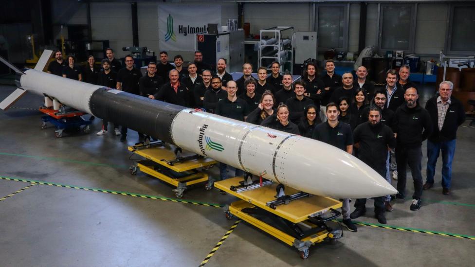 A long and thin white and black rocket lying on its side. A group of about thirty workers are gathered to the right. They're smiling at the camera and standing inside a warehouse.
