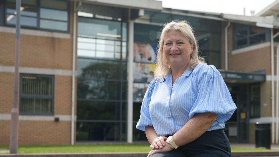 Bernadette Pettman is seen seated on a stone bench outside the modern school building, wearing a light blue striped blouse and dark trousers. Large glass windows and brick walls form the background, with a parked car visible to one side.