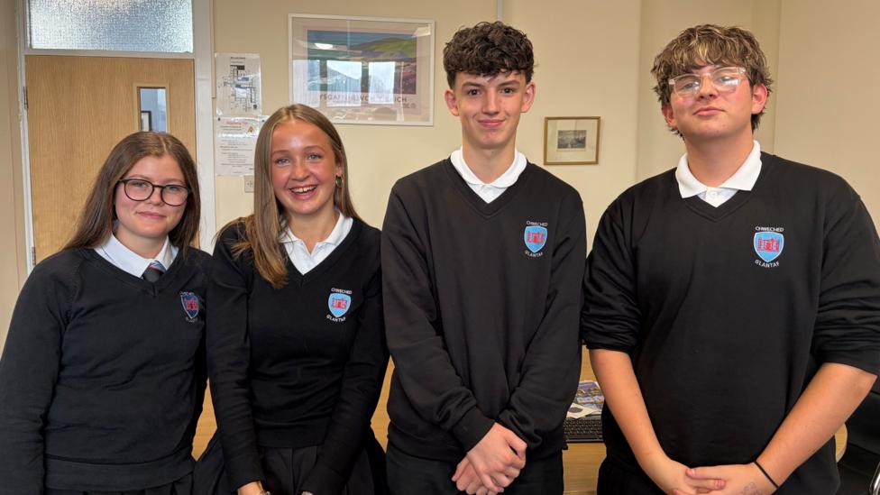 Four pupils in school uniform with two girls on the left and two boys on the right wearing dark jumpers and white shirts