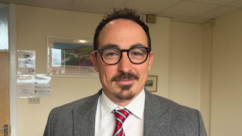A man with dark hair glasses and a beard in a grey jacket, white shirt and diagonal striped red and white tie.