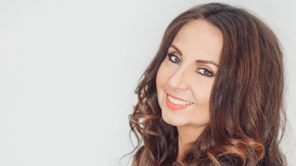 Melissa Alcruz with long brown wavy hair smiles at the camera, against a light background