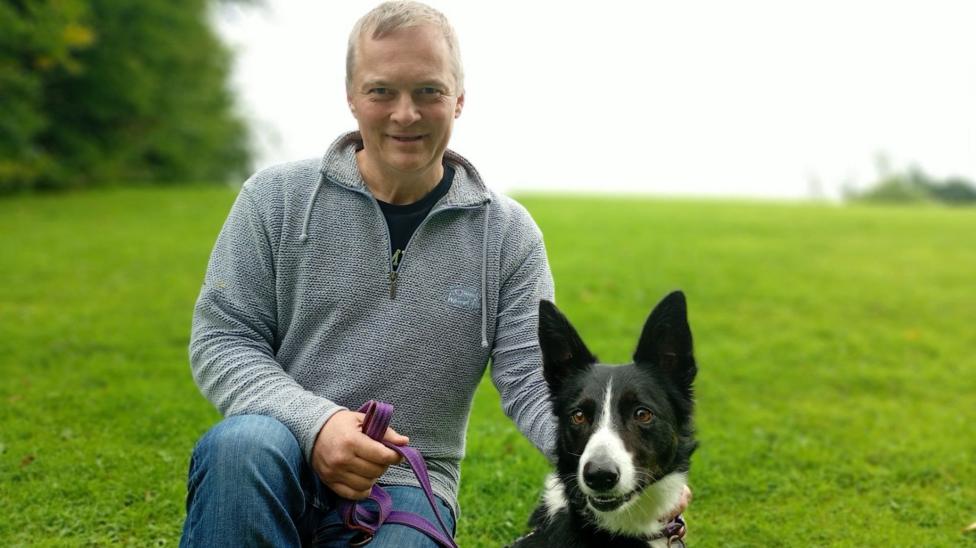 A man with grey hair wearing a grey 1/4 zip and jeans. He has a black and white dog and is holding a purple lead