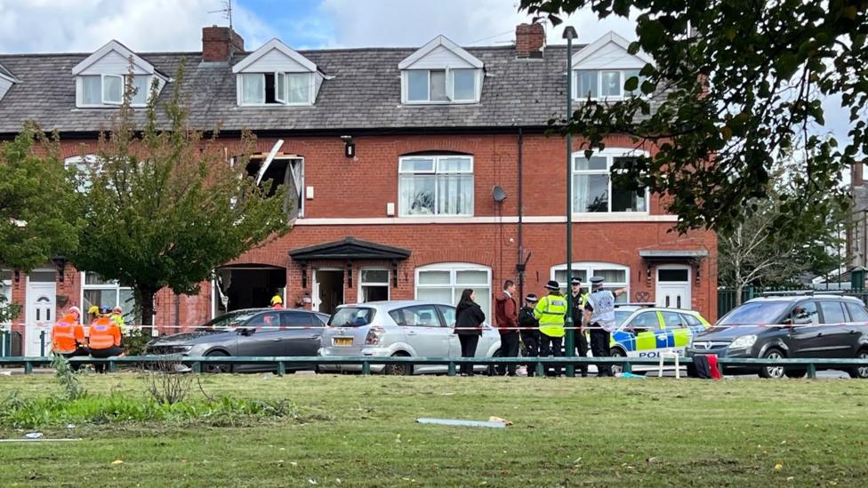 Image shows police and civilians standing behind a line of police tape, with a house which has had its windows blown out in the background. 