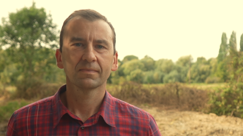 A head and shoulders image of a man wearing a shirt while standing in front of some trees. He is looking into the camera.