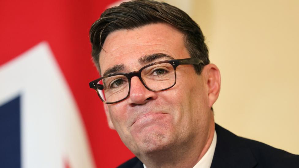 Andy Burnham smiling, in front of a Union Jack flag which is out of focus in the background. He is wearing a black suit, white shirt and dark rimmed glasses.