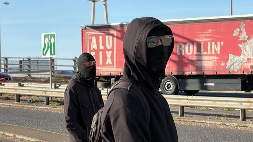 Two men wearing black clothing and black balaclavas. They are stood in front of a red lorry.