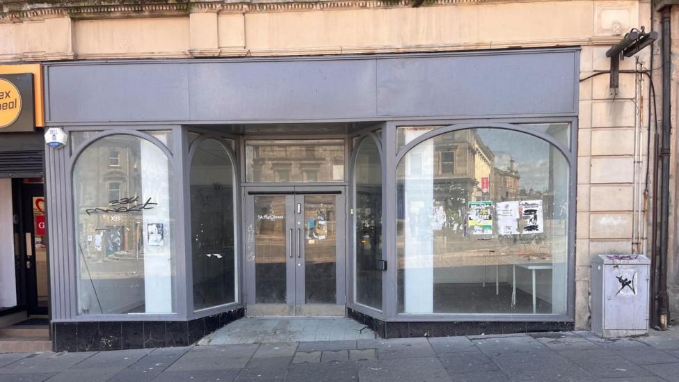 Vacant shopfront on a high street with large, curved display windows and double glass doors at the entrance. The exterior is painted grey, with empty interior space visible through the windows. Reflections of nearby buildings and street posters appear in the glass, while a pavement, utility box, and sections of neighbouring businesses frame the storefront.