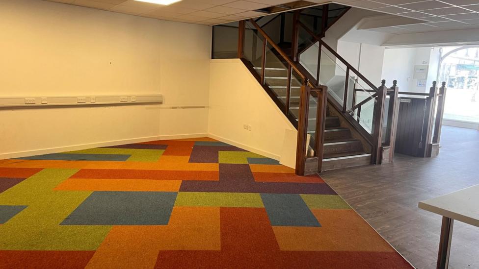Empty commercial interior with a colourful geometric-patterned carpet in shades of orange, red, green and blue. A dark wood staircase with glass-panel railings leads to an upper floor on the right side of the space. White walls, suspended ceiling tiles and a section of wood-effect flooring contrast with the bright carpet, while large front windows at the far end allow natural light into the vacant unit.