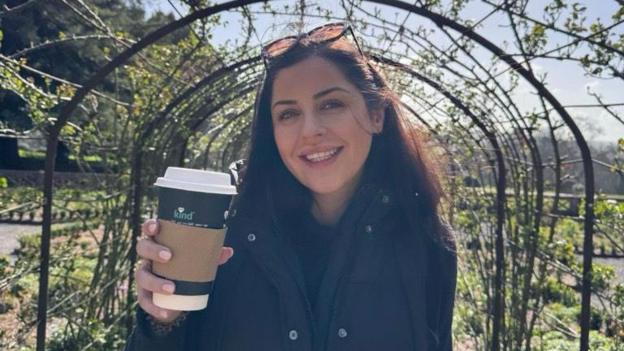 Erin Atkinson holds a takeaway coffee cup as she stands beneath a garden archway covered with climbing plants.