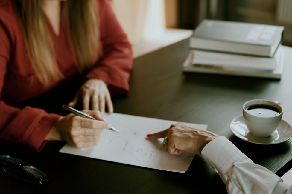 Two people sit at a table looking at a sheet of paper, with one pointing at the page and the other holding a pen. Books and a cup of coffee are visible in the background.