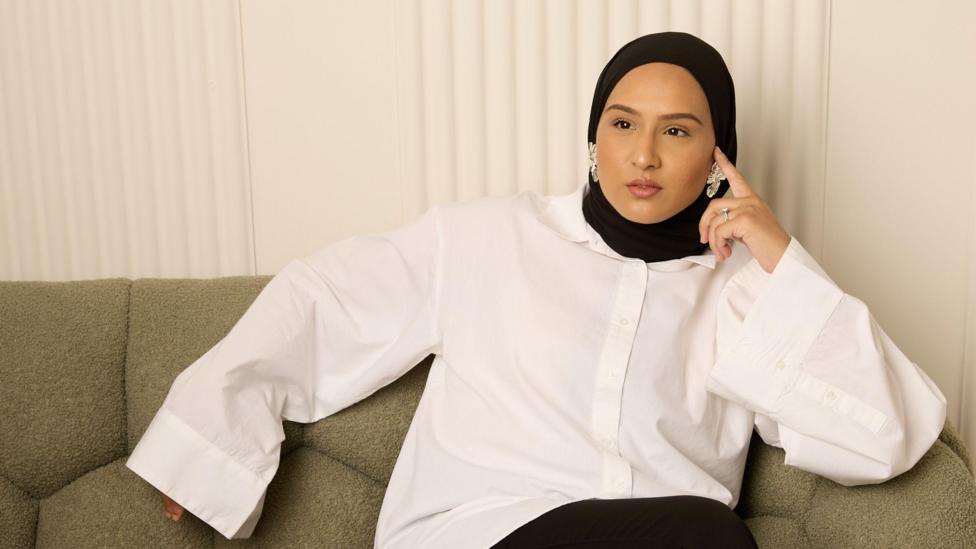 Tina Rahman looks thoughtful while sitting on a grey sofa and wearing a baggy white shirt.