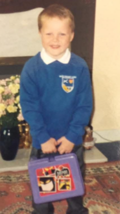 A young boy in an old film photograph, wearing a blue school uniform. He is smiling at the camera and holding a purple Batman lunchbox.
