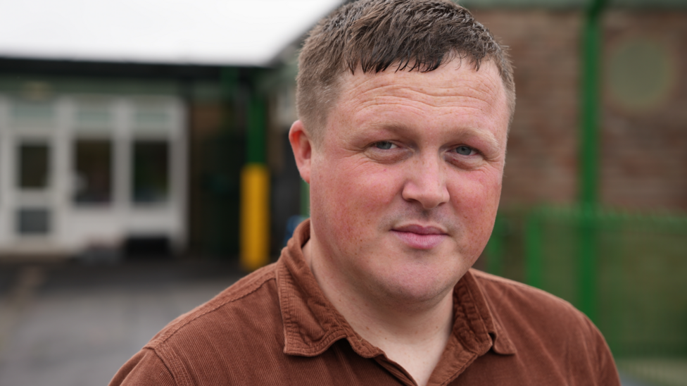 Male faces the camera with a neutral expression. He has short brown hair and blue eyes. He is wearing a brown shirt with a collar.