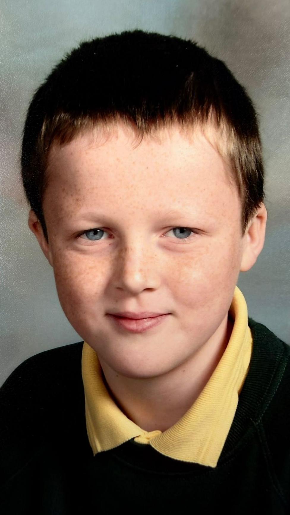 An image of a school picture of a white boy with brown hair wearing a dark jumper and a yellow t-shirt