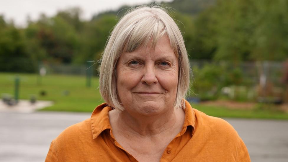 An image of a white woman with short blonde hair wearing a dark yellow shirt with a school field and playground in the background