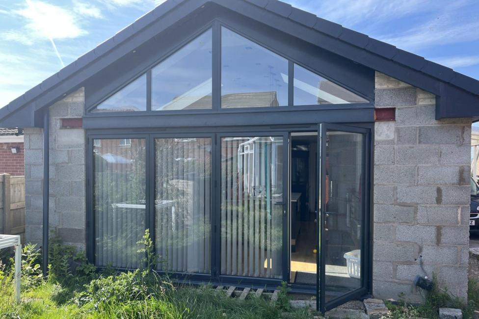 The rear of a bungalow with a pitched cathedral roof and grey bi-fold doors with vertical blinds, taken from outside. The walls are grey breeze block and un-rendered, the grass on the lawn is unkempt and there is building rubble outside the doors. One of the doors is open and the kitchen floor is just visible inside as well as the edge of a kitchen island.