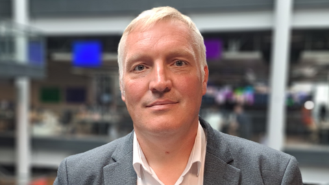 A white man with white hair wearing a white shirt and grey suit stands in front of an office background