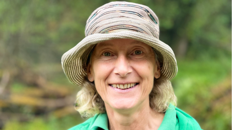 Kate Wollen from Forestry England smiles at the camera, she is wearing a round brimmed hat and stands in a forested area