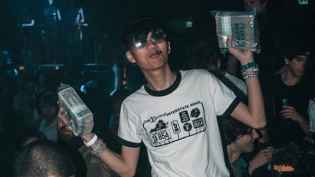 Malone Lam wears a T-shirt and holds up stacks of cash in a nightclub