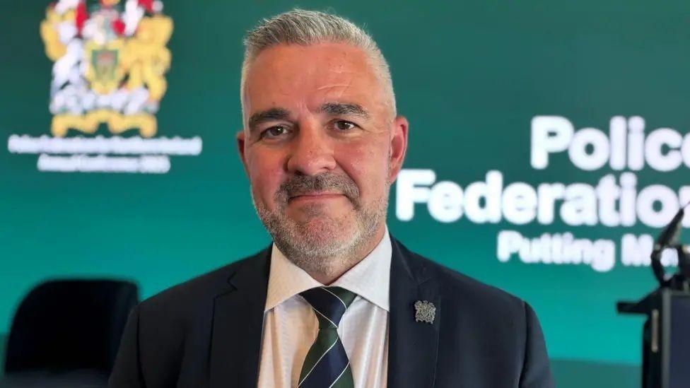 Liam Kelly has short grey hair and facial hair. He is standing in front of a green screen and is wearing a black suit jacket, white shirt and navy and green striped tie.
