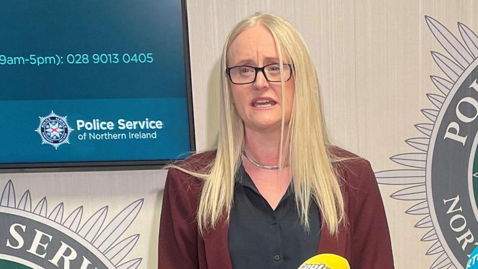 Geraldine Hanna speaking into microphones at a media conference. She is wearing a black blouse and burgundy jacket. She has long blonde hair and is wearing glasses.